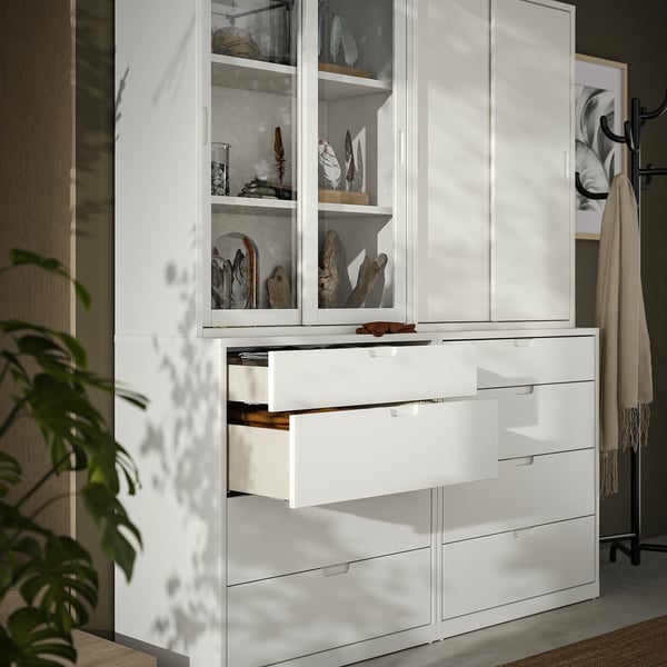 White TONSTAD cabinet and chest of drawers with glass doors and wooden handles, displaying decorative items, next to a towel rack and a plant.