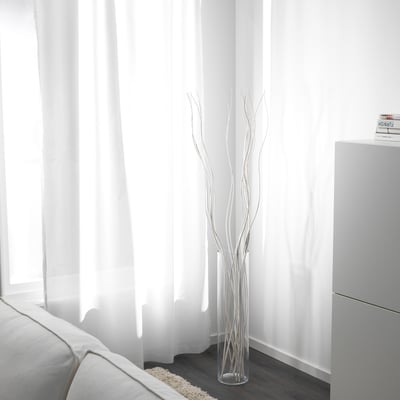 Clear vase with white dried twigs near a window with white curtains and a partially visible white couch.