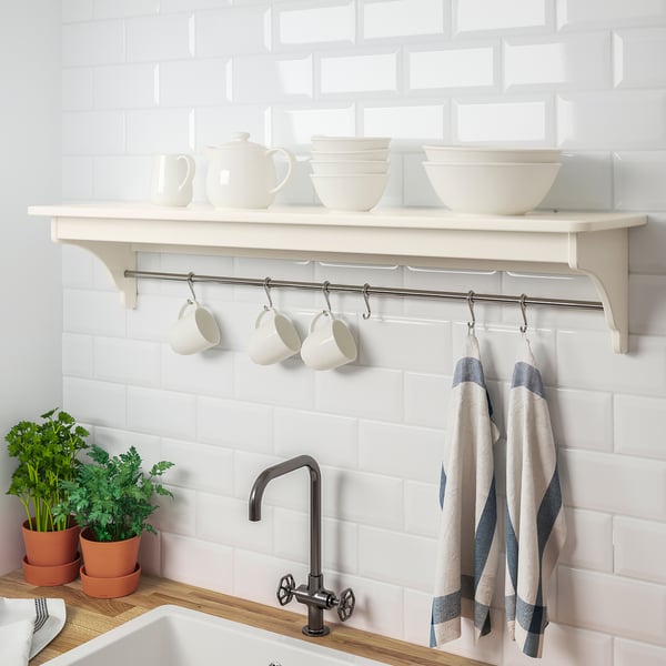 A white kitchen shelf holds white crockery above mugs and striped towels, mounted on a tiled wall.