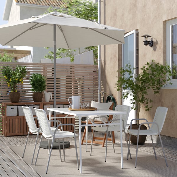 Modern outdoor dining set with white chairs and table, large beige umbrella, potted plants, wooden fencing, and beige building in background.