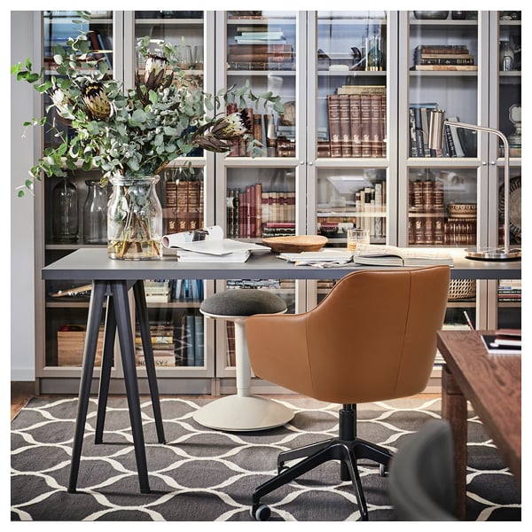 Home office setup featuring TOSSBERG brown leather swivel chair, grey desk, and glass cabinet filled with books. Patterned rug; cosy, modern design.