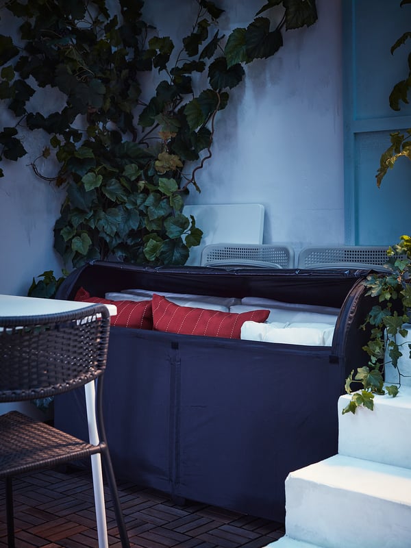 Dark blue waterproof TOSTERÖ storage box, outdoor cushions inside, red pillows, wicker chair, white wall, ivy.