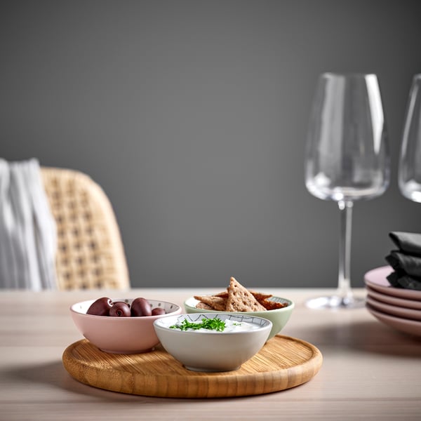 Three colourful bowls on a bamboo tray hold olives, dip, and crackers. Wine glasses and plates nearby suggest a shared meal.