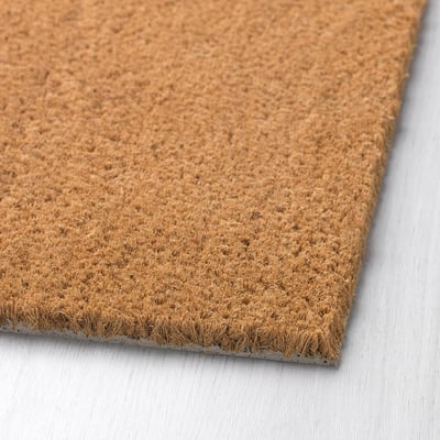 Close-up of textured coir door mat on wooden floor. Natural fibres and latex backing visible. Clean, rustic appearance.
