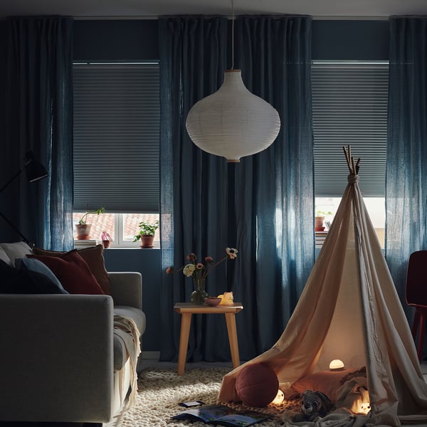 Cosy living room with dark curtains, a lit teepee tent for kids, and grey sofa. A hanging lamp provides soft light.