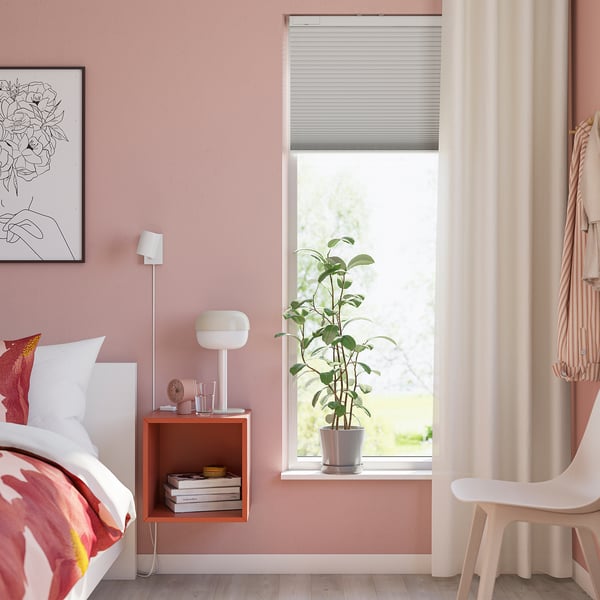 Cosy bedroom with floral bedding, pink walls, TREDANSEN blind, plant, and side table with lamp and books.