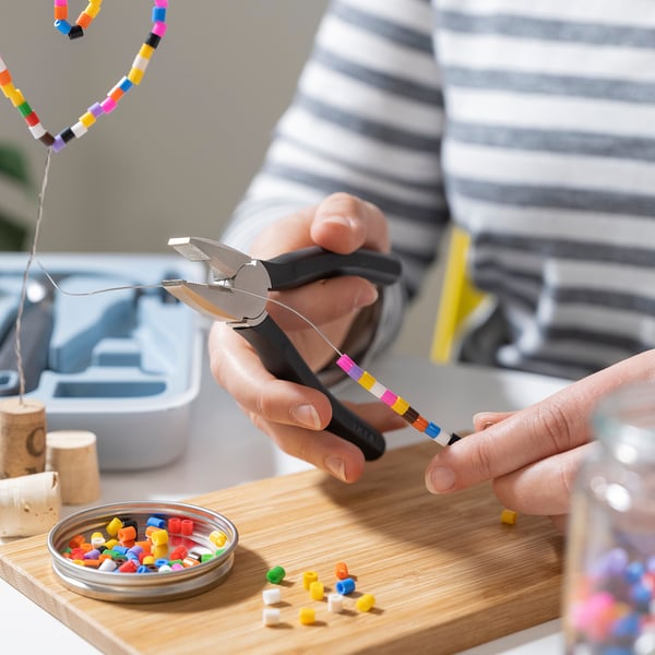 Hands use TRIXIG pliers to bend wire with colourful beads. Tools and beads on wooden board for diy crafting.