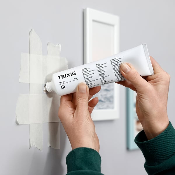 Person squeezes white TRIXIG repair filler onto wall patch. Product label shows details and multi-language instructions.
