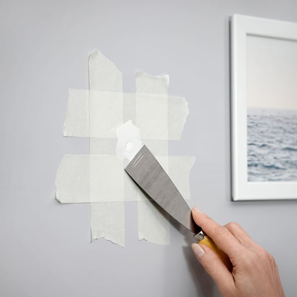 Person repairs a wall hole using putty knife and paper tape. Wall has grey paint, and a framed picture of ocean is beside.