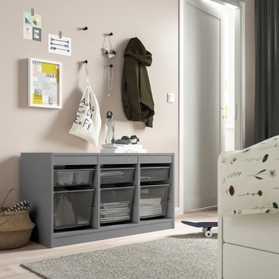 Grey TROFAST storage unit with mesh baskets, hanging jackets, wall art, near a skateboard and cot.