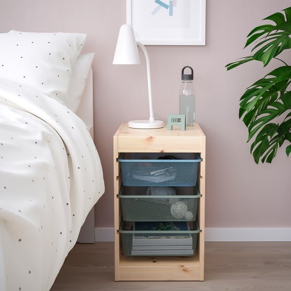 Wooden TROFAST unit beside bed with lamp, water bottle, and clock. Three drawers, white comforter on bed.