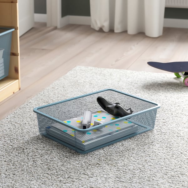Mesh storage basket on a carpeted floor, holding a game controller and a toy. Part of the TROFAST series, providing organised storage for kids items.