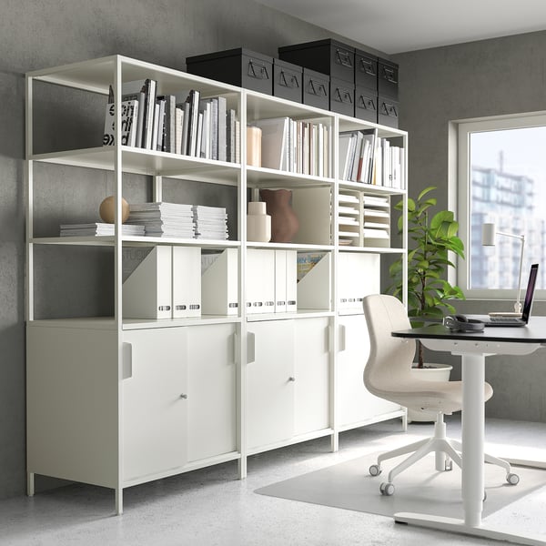 White TROTTEN shelving unit in grey room, holding books, boxes, and decor. Modern metal design, smaller than chair, next to a window.