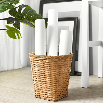 Wicker basket TRUMMIS holding white items, set on wood floor with plant and frames.