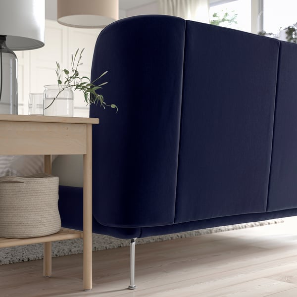 Modern bed with curved, dark blue headboard. Wooden legs and storage space underneath. Decorative plant on the wooden side table next to it.