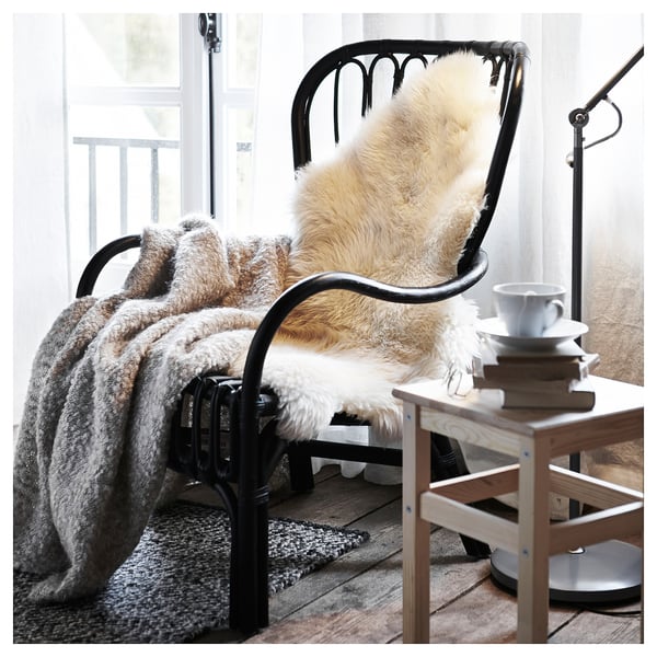 Black metal chair with beige fur throw, grey fur blanket, cup, books, and lamp on a wooden side table near a window with sheer curtains.