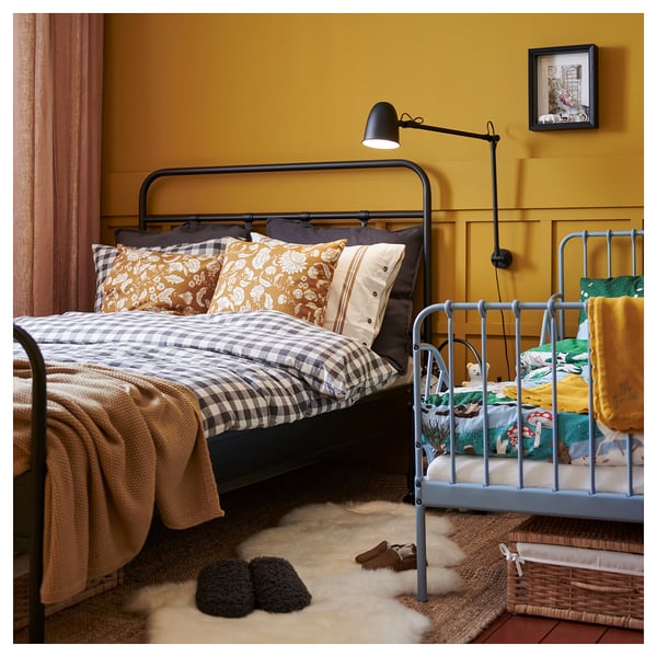 Cosy bedroom with black bed, yellow pillows, blue kids bed, beige rug, slippers, wall sconce, framed picture on yellow walls.