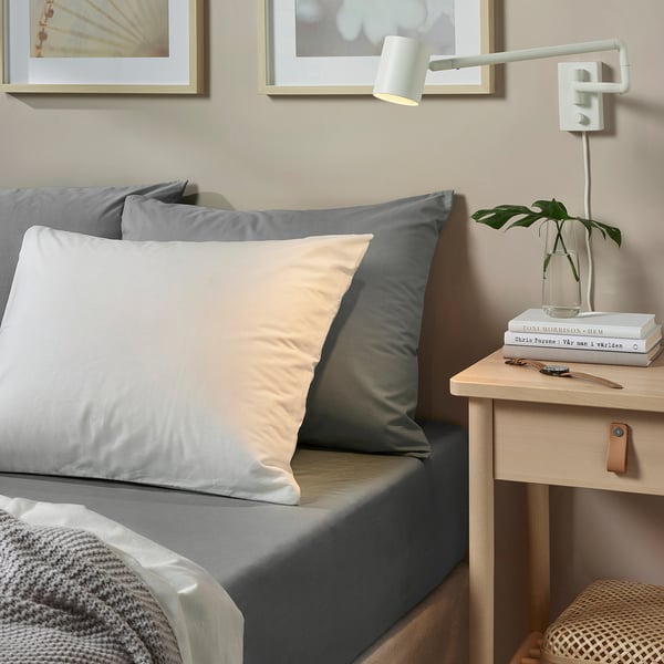 Neutral bedside with grey & peach pillows, a cosy blanket, stacked books, and a wall lamp.