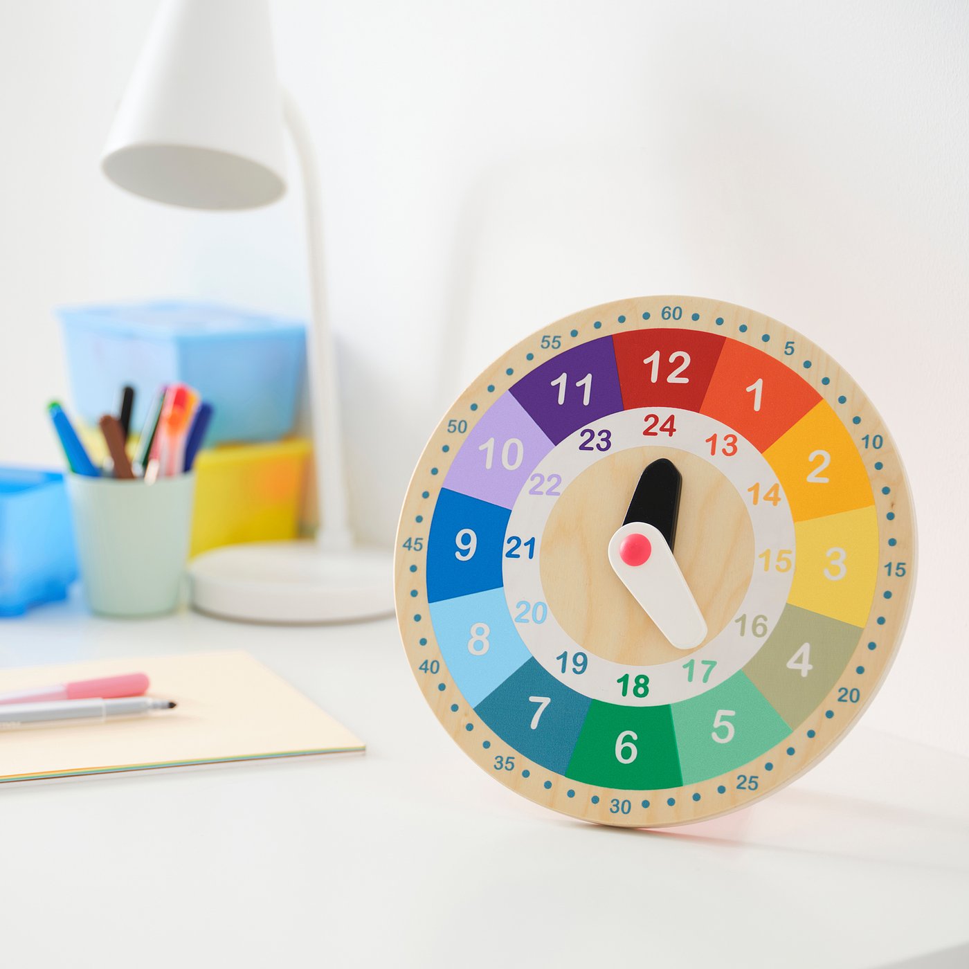 Educational clock on desk with stationery and lamp.