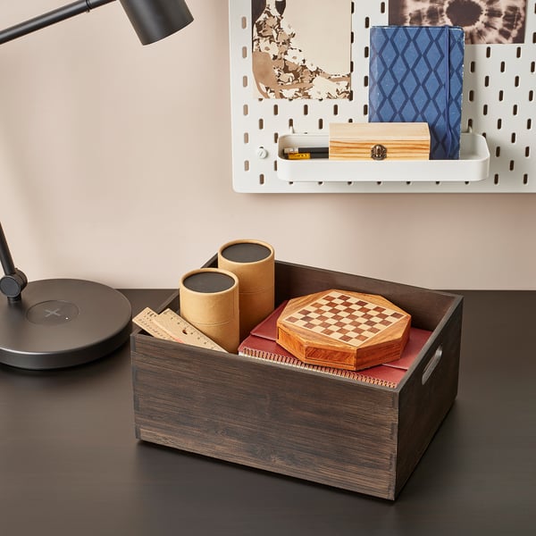 Wooden box on desk holding two cylindrical containers and a chess set. Beside a lamp and white storage with books and items.