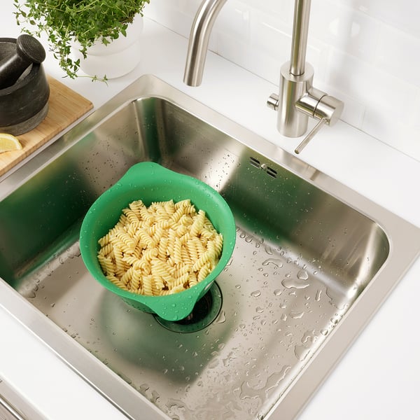 Stainless steel kitchen sink with green colander full of cooked pasta, placed on left side. Silver tap on right.