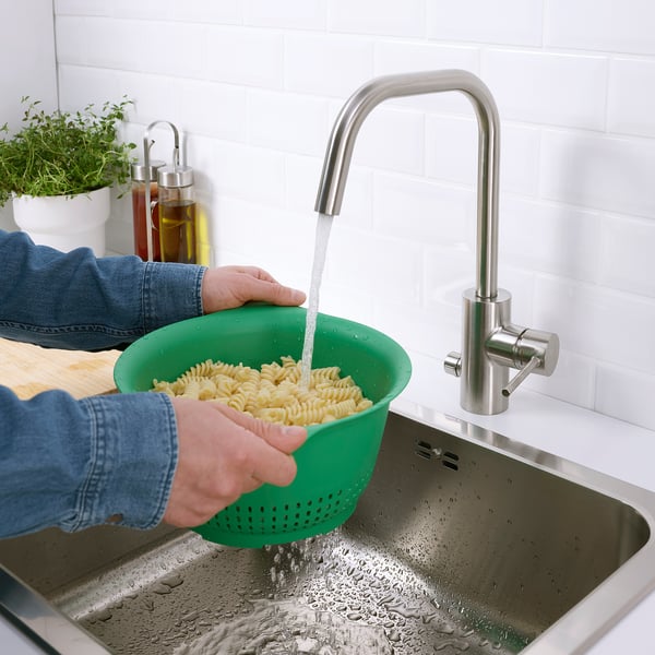 Person rinsing cooked pasta in green UPPFYLLD colander under stainless steel tap.