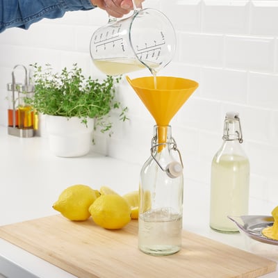 Person pours liquid from measuring cup through yellow funnel into glass bottle. Lemons, another bottle, and herbs nearby.