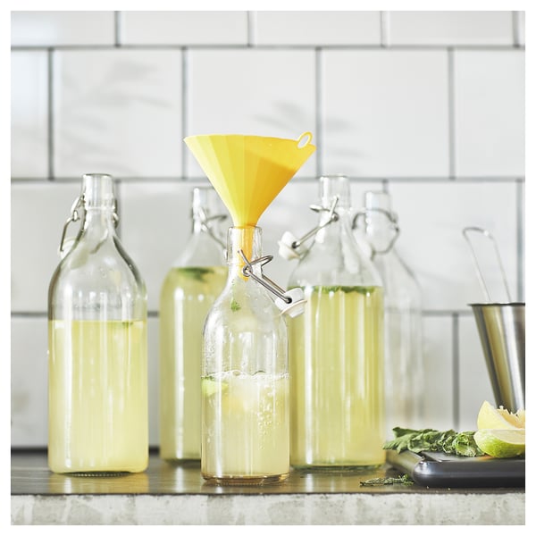 Four glass bottles with yellow liquid and a yellow funnel. Against a tiled wall. Leftover lime and mint on a cutting board.