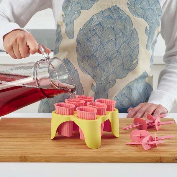Person pouring red liquid into pink silicone ice pop moulds on a wooden board. Moulds are part of the UPPFYLLD brand, used to make fruit ice pops.