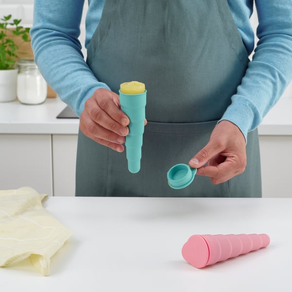 Person holding ice pop mould, demonstrating how to assemble it. Other moulds on kitchen counter, alongside cloth and jar.