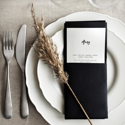 Black menu card on white ruffled edge plate, silver fork and knife, dried plant stem, grey tablecloth.