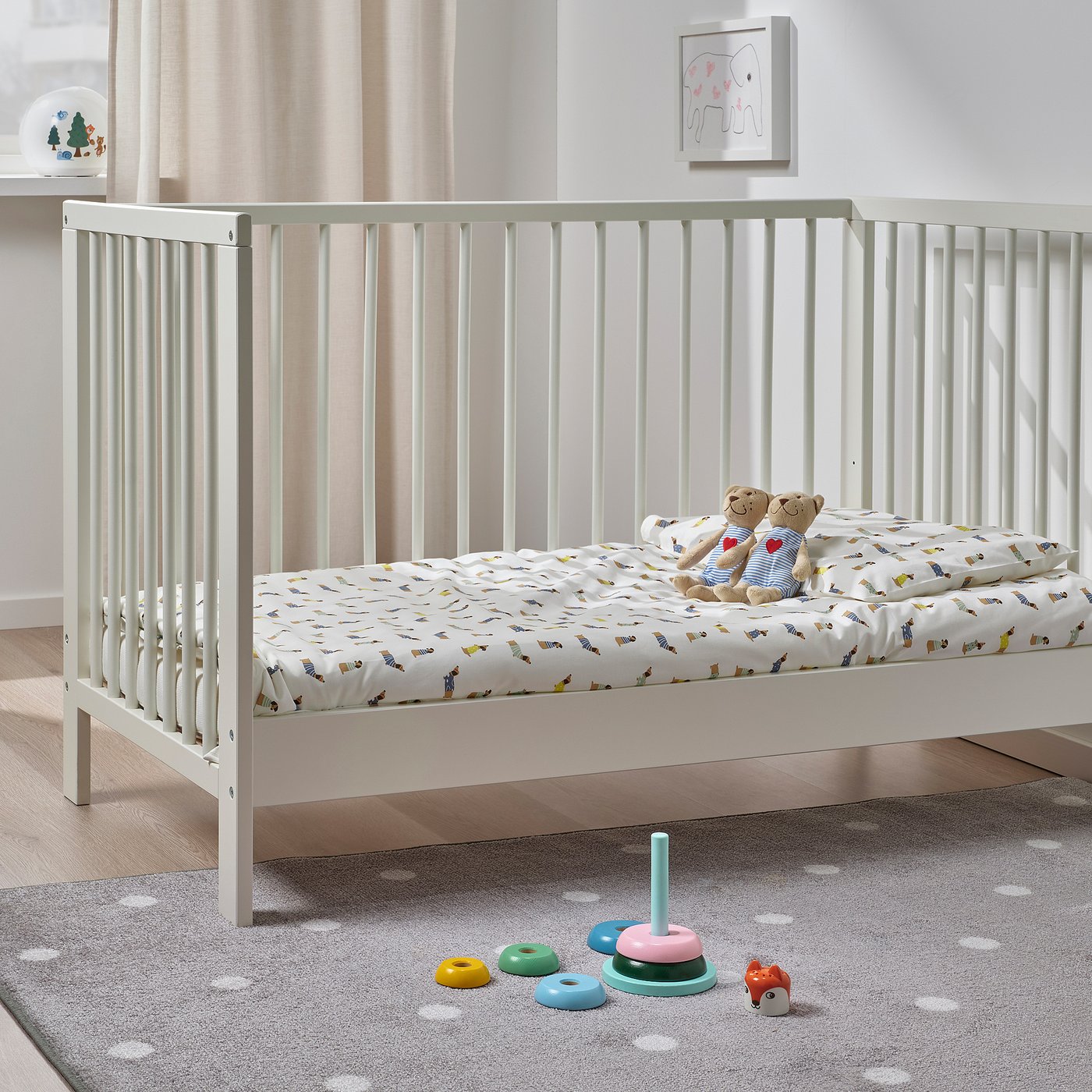 Baby crib with teddy bears, colorful stacking toy, and polka dot rug. Artwork on wall.