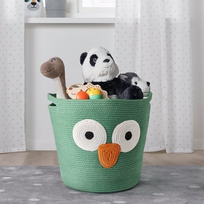 Green cotton basket with owl design holds plush toys on grey polka dot rug.