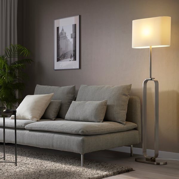 An UPPVIND floor lamp with a slim, oval shade, standing tall beside a grey sofa, casting a warm glow.