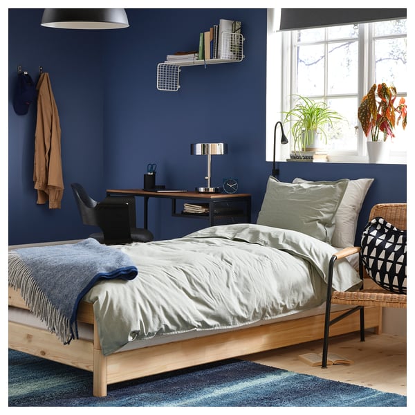 A modern blue bedroom with a wooden bed frame, white bedding, a black desk, and a window with plants.