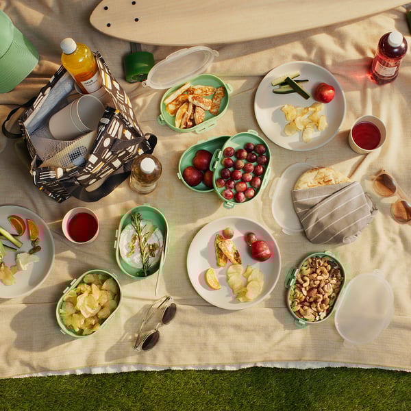 Picnic setup with sandwiches, fruits, nuts, and drinks on a blanket. Includes green stackable lunch boxes and a patterned cooler bag.