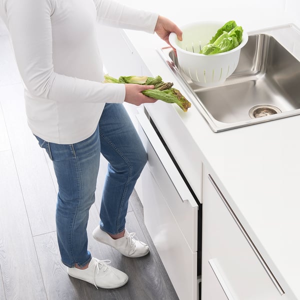Person in white shirt and jeans uses a kitchen drawer with a push opener, holding green lettuce over a sink.