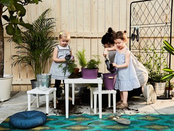 Two kids playing with UTTER white childrens table and purple buckets, surrounded by plants and garden tools.