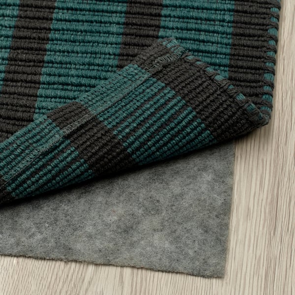 A dark green and black striped rug, positioned on a grey rug pad on a wooden floor. The rug has a flat surface, making vacuuming easy.