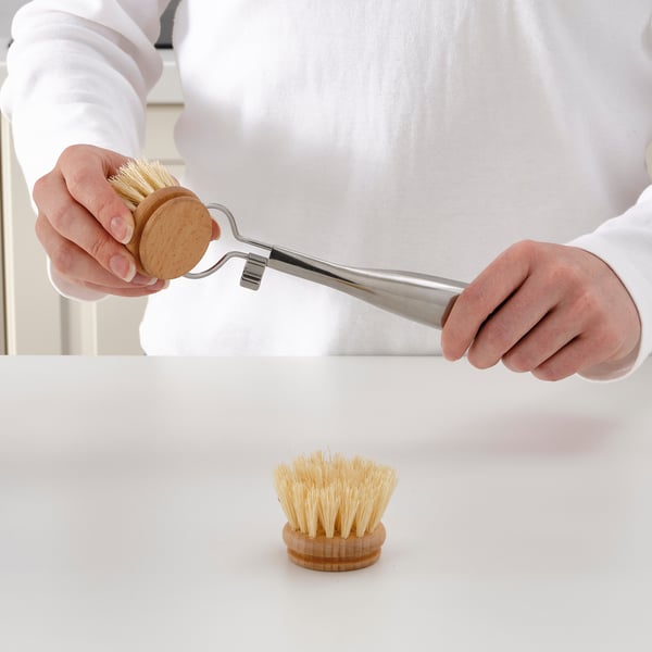 Person holds a dish-washing brush attachment with a metal handle, demonstrating its use. The brush head can be detached and replaced.