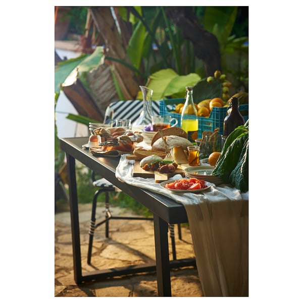 Outdoor meal setup with sandwiches, fruits, and drinks on a black VÄRMANSÖ table surrounded by greenery.