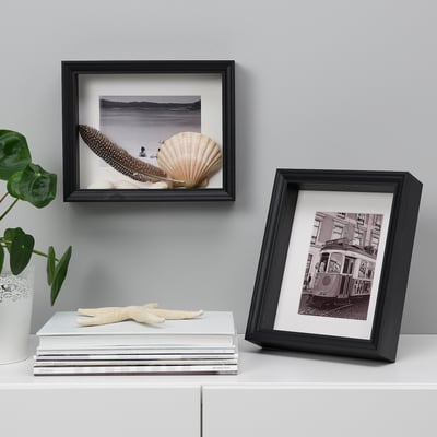 Two framed artworks: seashells & feathers, tram. Stack of books, starfish, plant on white surface against grey wall.