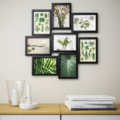 Wall-mounted collage of eight framed green plant illustrations, part of IKEAs VÄXBO series, arranged above a shelf holding candles and books.
