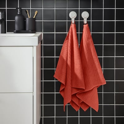 Two bright orange VÅGSJÖN towels hang on wall hooks beside a modern sink with dark tiles and black bathroom accessories.