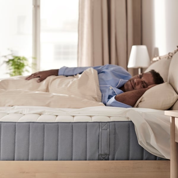 A person sleeping on a blue VALEVÅG mattress on a wooden bed frame. The white bedding and lamp are also visible.