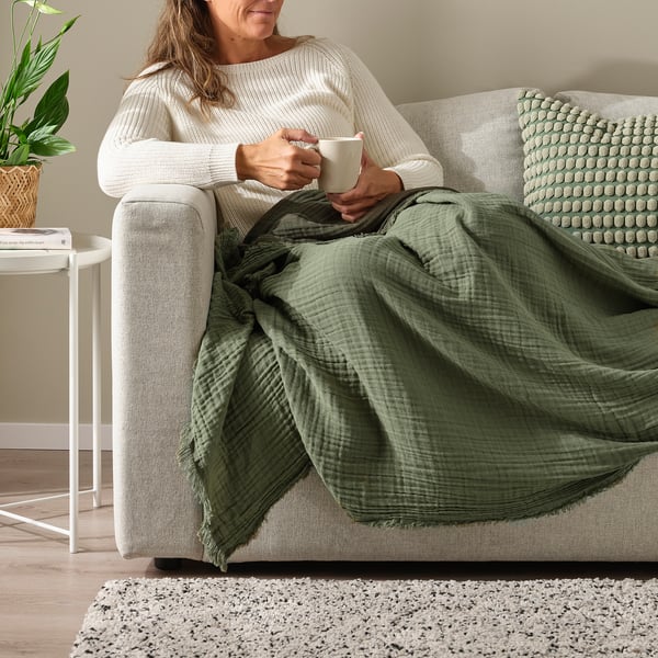 A person relaxes on a beige sofa, holding a cup of coffee. Shes wrapped in a green VALLKRASSING throw blanket.