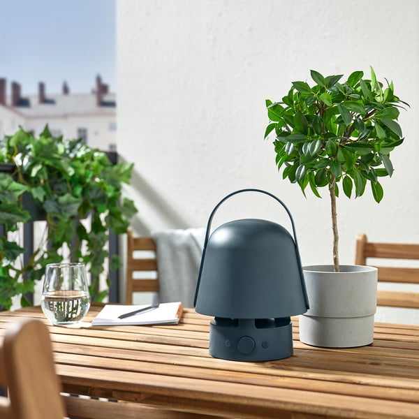 Outdoor table with VAPPEBY blue speaker-lamp, small potted plant, glass, and notepad.