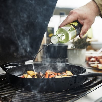 Person cooking with VARDAGEN black cast iron skillet, pouring oil, smoke rises, outdoor setting.