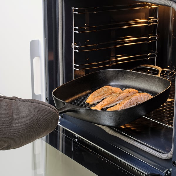 Image shows a person placing a cast iron pan with cooking salmon into an oven, highlighting the pans oven-safe feature.