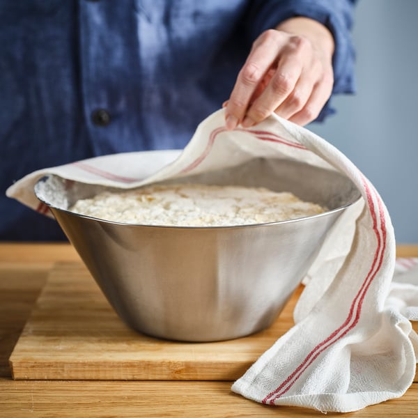 Person covering a large stainless steel bowl with a cloth. The bowl has a spout and sits on a wooden board.